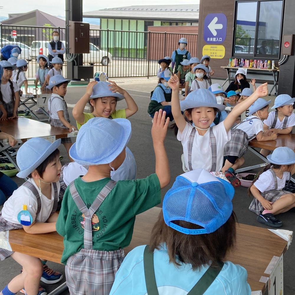 手前の机の周りに並んでいる園児達が手をあげ喜んでいる様子を写した写真