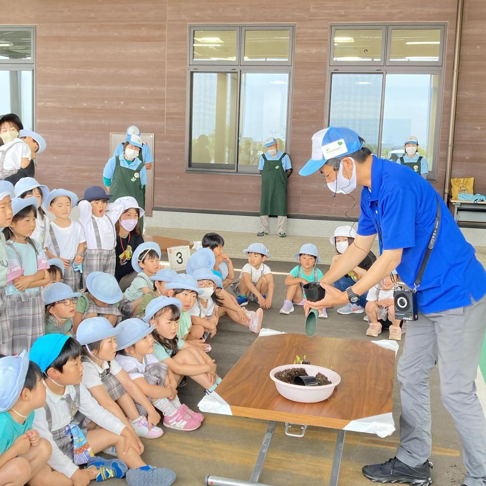 机の上に置いたおけからポットに土をいれる説明を園児達が座って聞いている写真