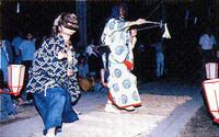 夜の薄暗い中で、面をかぶった和装の演者が二人踊っている様子の写真