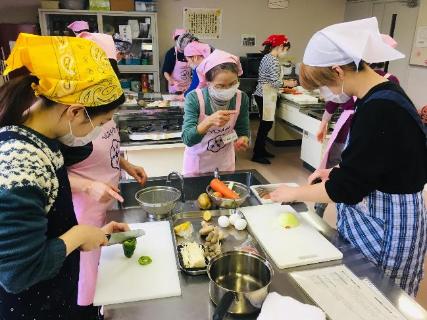 三角巾とエプロンを付けた参加者の方々がグループに分かれて調理台で野菜を切っている養成講座の調理実習の様子の写真