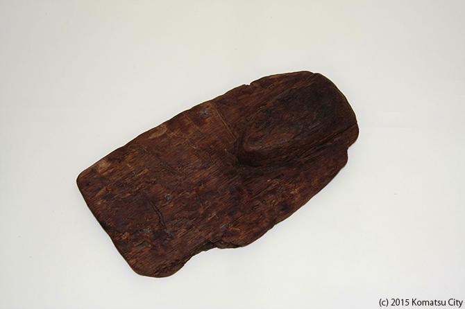 長方形の片方の先が丸くなった形の濃い茶色の木の板で、角のある端部が欠けている鍬18の写真