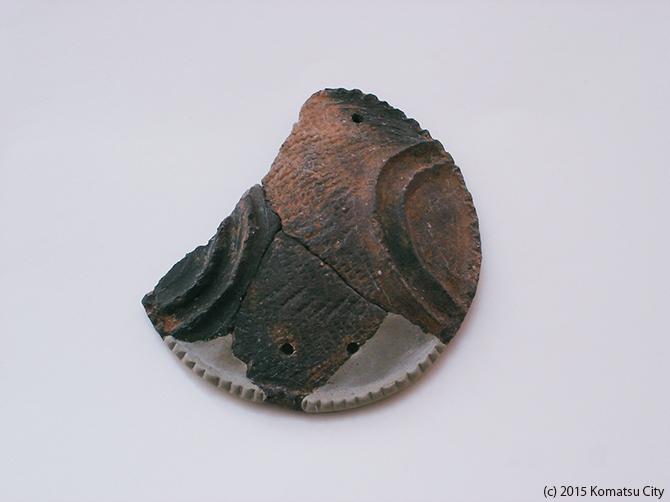 側面がギザギザで上面が半分に分かれ右はこげ茶色に黒、左が黒に灰色で突き合わされ、穴が3ケ所空いている蓋形土器1通番号:140の写真
