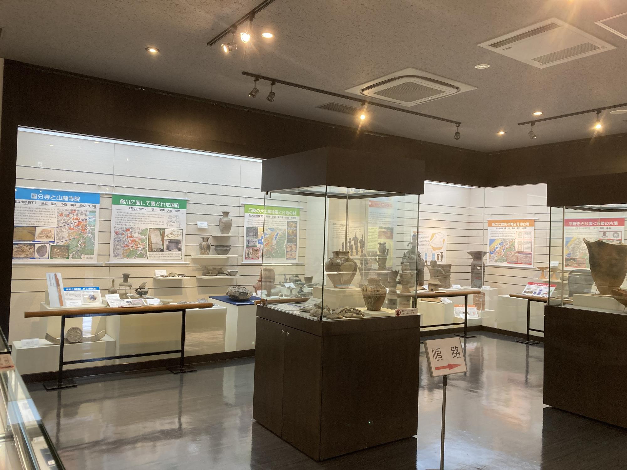 壁などに設置された展示ケースに、説明とともに出土品の土器などが並んでおり、室内には順路を示す看板が置かれている埋蔵文化財センター内の写真