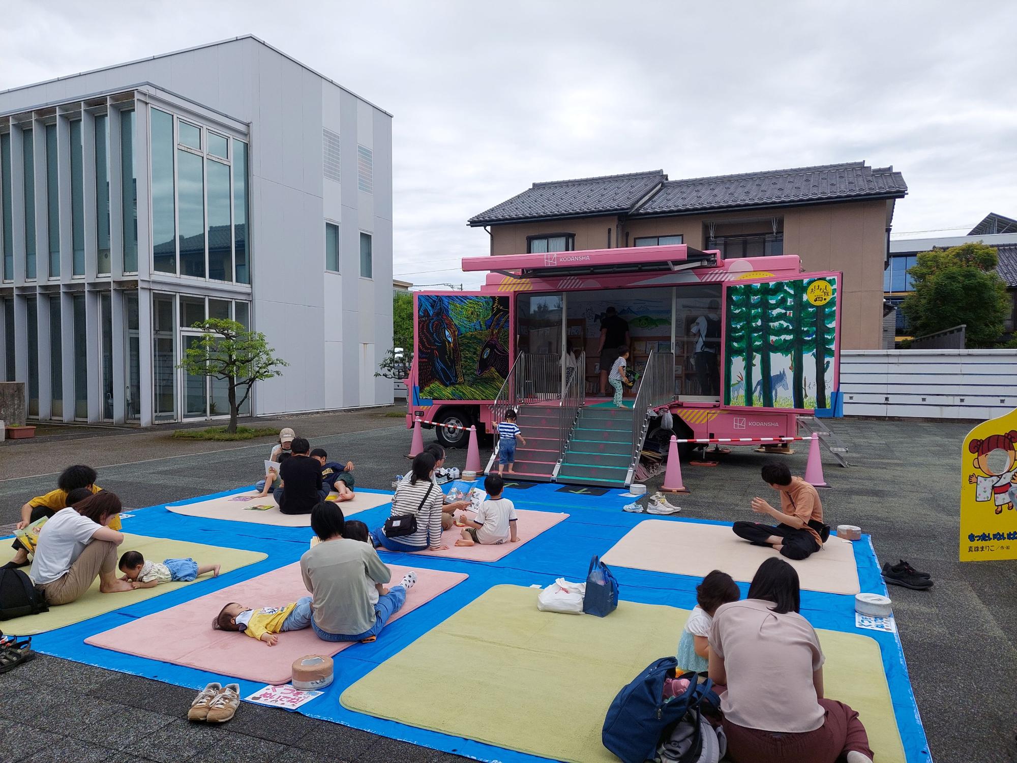 絵本館広場に敷かれたマットの上で絵本を読んでいる親子の画像