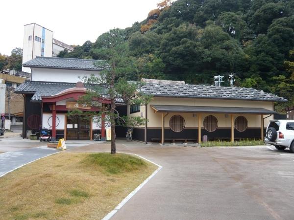白壁で瓦屋根をもち、入口部分に赤い門と「総湯」と書かれた店名板が設置された左側の建物と、ベージュを基調とし下部分が黒、丸格子の窓をもつ右側の建物が併設されている粟津温泉総湯の外観写真