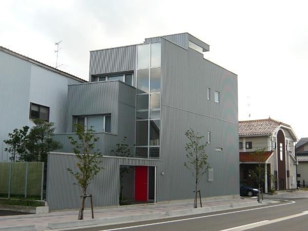グレーを基調とした3階建ての建物であり、一部がガラス壁の仕様、前庭には樹木が植えられている、赤の玄関ドアが目を引くモダンで清楚な高橋邸の写真