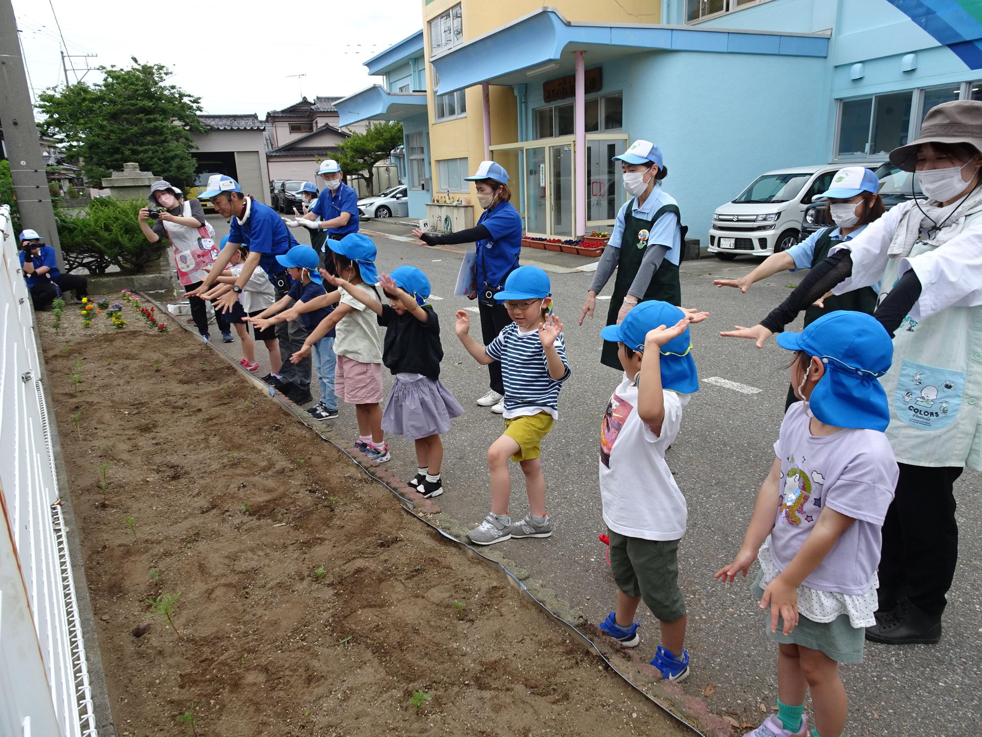 花の苗を植え終わった花壇の傍で一列に並んだ園児たちが、花壇に向かって両手をかざしている様子の写真