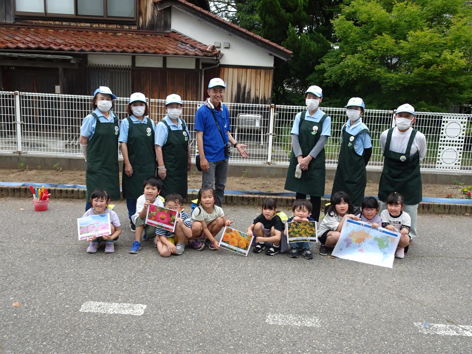 4種類の花の写真と世界地図を持った9名の園児と、6名のスタッフと寺井先生が並んでいる集合写真