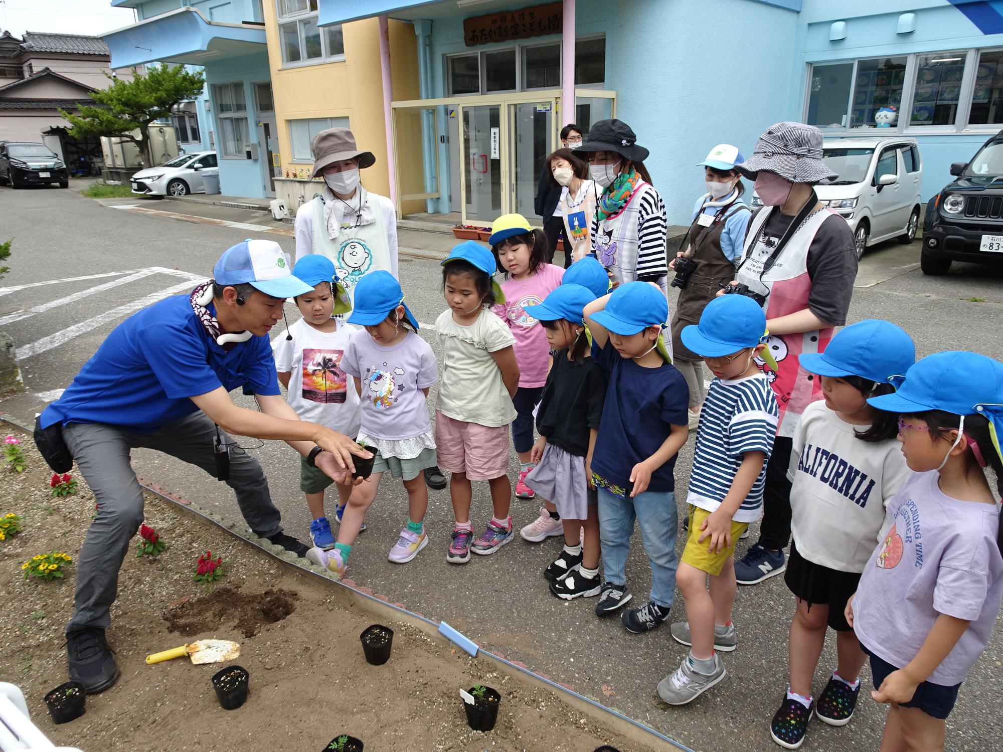 駐車スペースがあるベージュ色と水色の外壁の建物の手前の花壇で、苗ポットを持って説明を行っている寺井先生の話を園児たちが聞いている写真