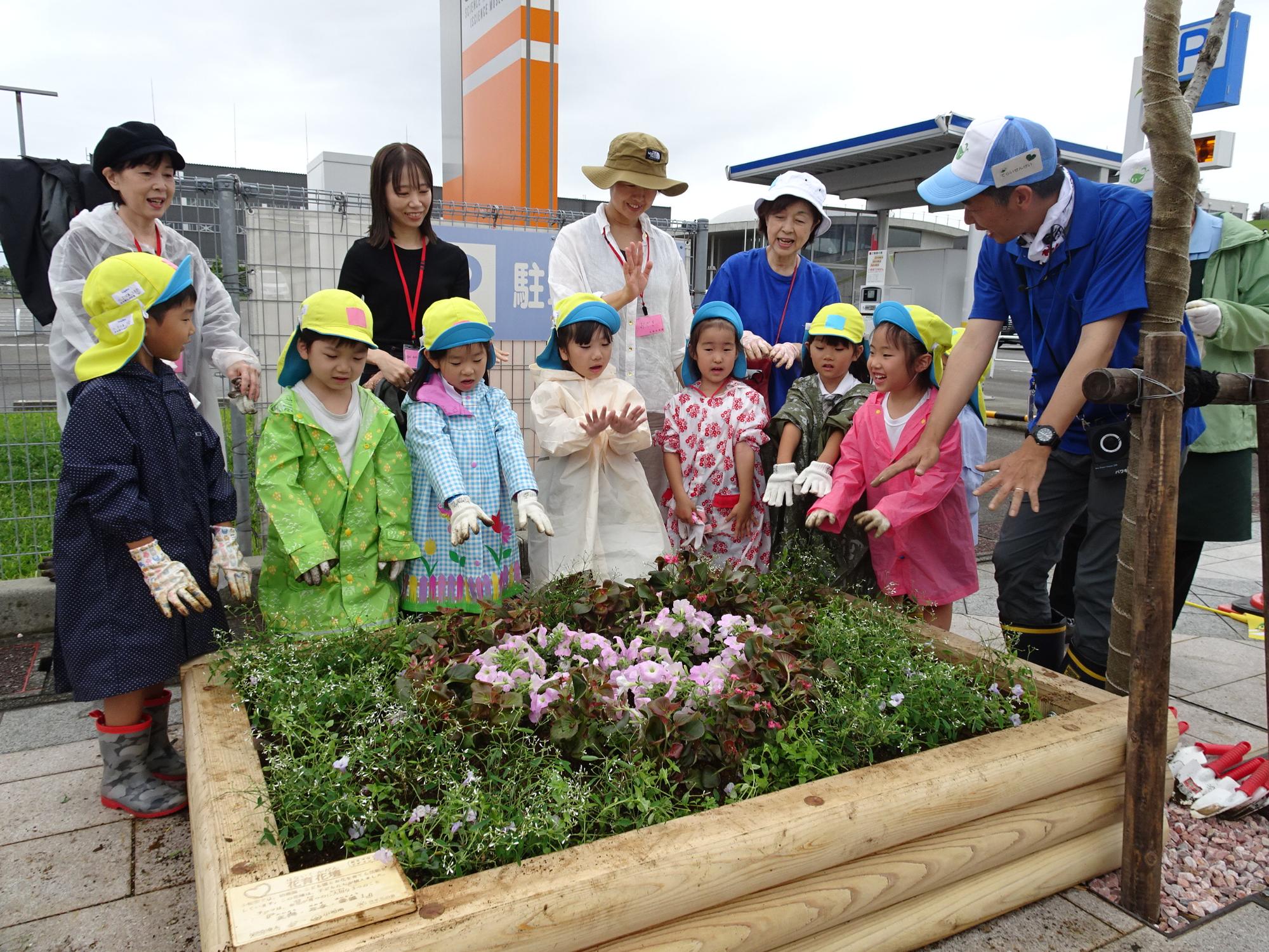 花壇の周りに7名の園児と4名の保護者の方々、寺井先生が立って花を植えた花壇に両手をかざしている様子の写真