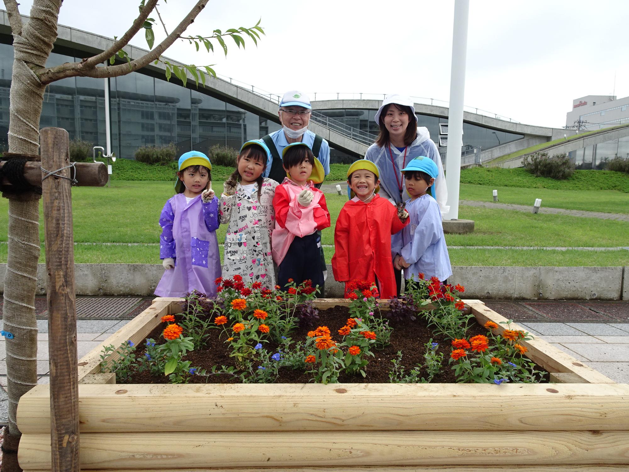 5名の園児と1名の保護者、男性スタッフが赤色やオレンジ、青色の花が植えてある花壇の後ろに並んでいる写真