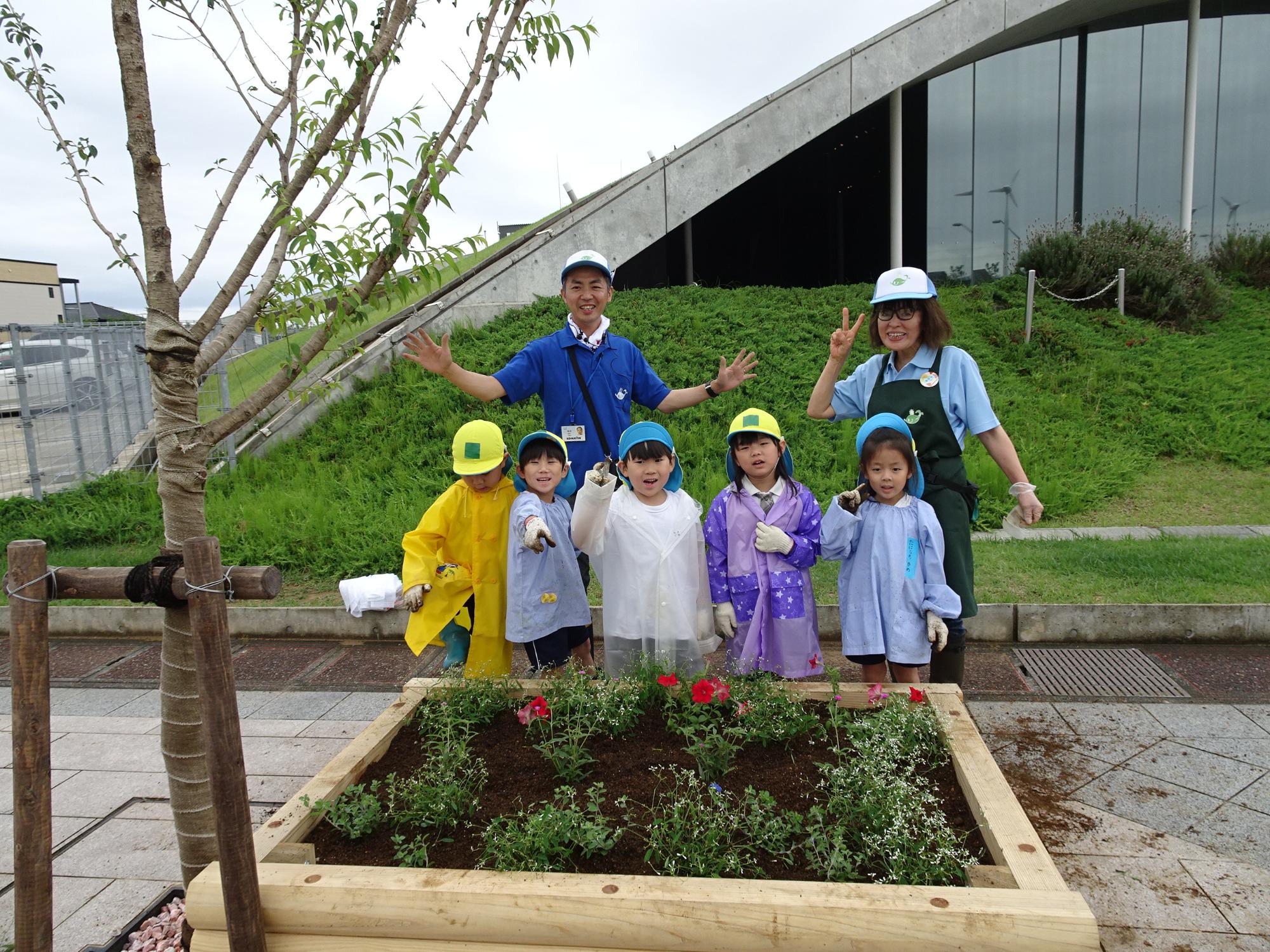 5名の園児と寺井先生、女性スタッフが赤色やピンク色の花が植えられた花壇の後ろに並んでそれぞれポーズをとっている写真