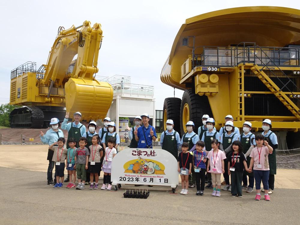 黄色の重機のをバックに「こまつの杜 2023年6月1日」と書かれた看板の場所で記念撮影をしているさくら組の皆さんの集合写真