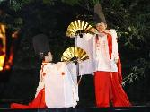 金色のセンスを持って巫女装束を着た2人が向かい合い、片方がしゃがんでいる原町の御前様祭りの様子の写真