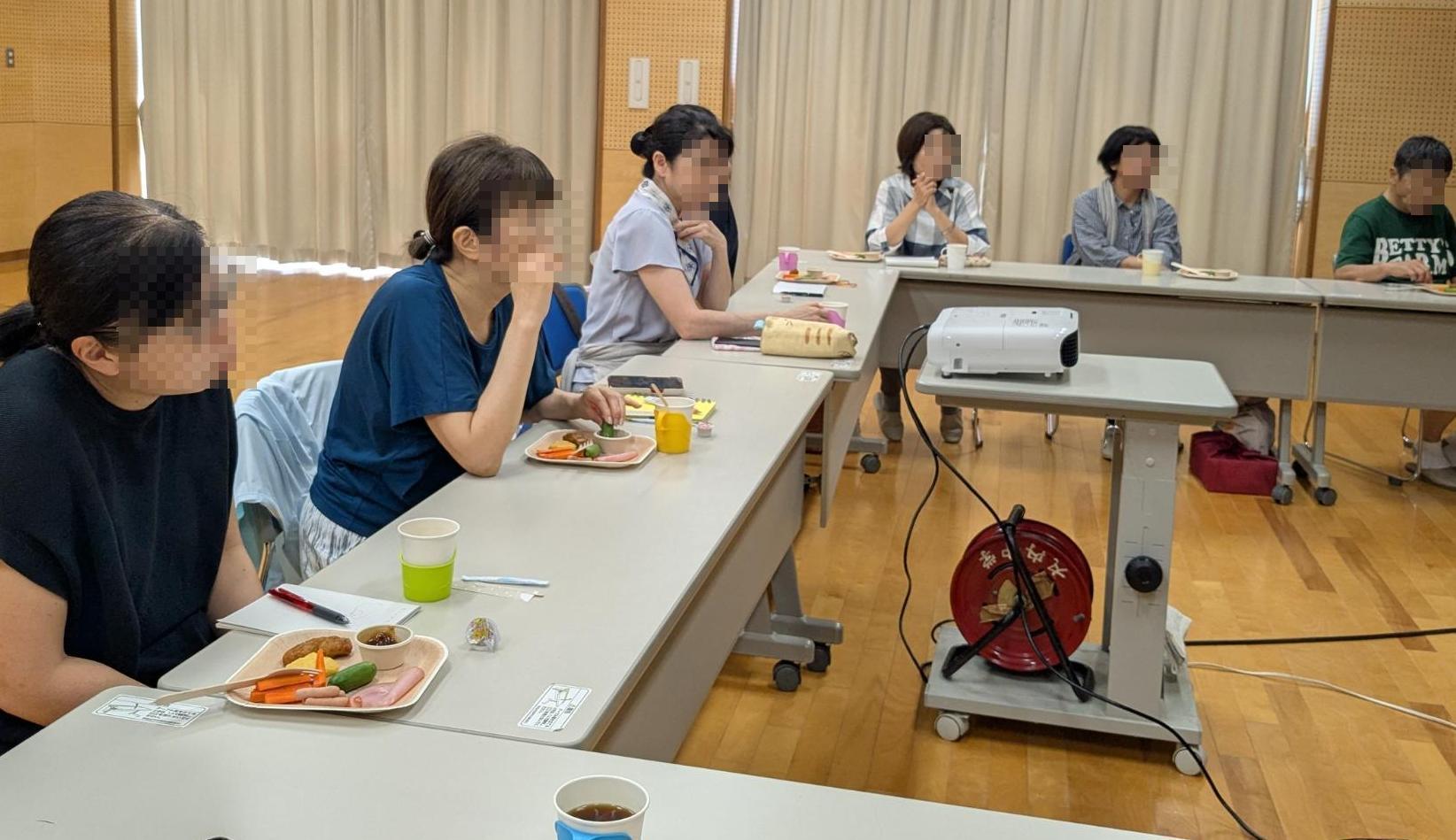 参加者6名が座って話を聞いている。机の上には食べ物がある。