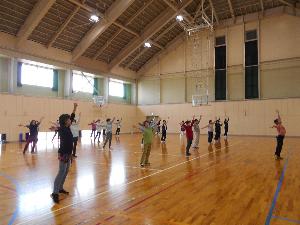 体育館の中で多数の参加者と講師が両手と両足を広げて体操をする様子