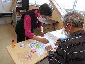 部屋の中で机の上に折り紙を並べて椅子に座っている男性の前に立ち指をさしている女性の様子