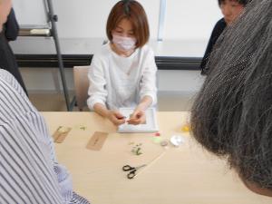 机に座った講師が紙の花びらを手に持ち糊をつけながら説明する様子