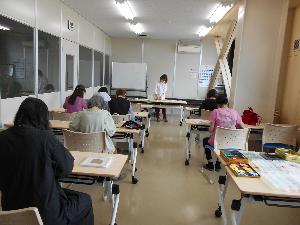 教室の中で7人の女性が椅子に座って机に向かいペーパーフラワーを作り奥に講師が立って下を向く様子
