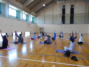 体育館の中で17名の女性がヨガマットの上で胡坐をかき左手を上げるポーズをする様子