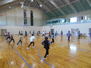 体育館の中で29名の女性が腰に手を当て右足を後ろに出し左足を曲げて前に出すポーズをとる様子