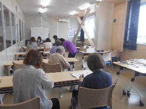 研修室の中で8名の受講生が長机に置いてある水引を使い講師の説明を聞きながら作品制作をする様子