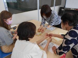 長机を2つ合わせた場所を3名の受講生が囲んで座り1人の女性が両手を出しながら水引作品を作るところを講師が手を添えて教える様子