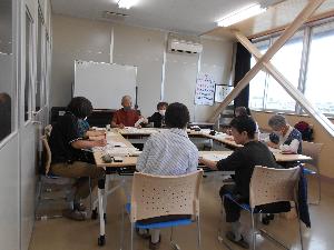 研修室内でテーブルを参加者7名と講師が囲み資料を見ながら講師の説明を聞く様子の写真