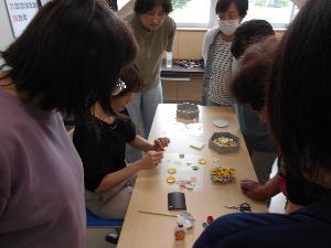 長机の前に座った講師を参加者8名が囲み机の上に置いてあるペーパーフラワーを組み合わせながら説明する講師の様子