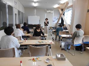 研修室の中で講師が部屋の奥に立ちながらペーパーフラワーを持ち8名の参加者が説明を聞く様子