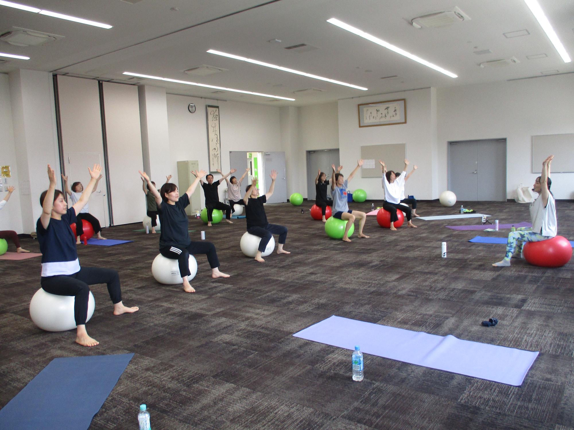 バランスボールエクササイズの様子、先生の指導をみながらバランスボールに載って両手をあげている。