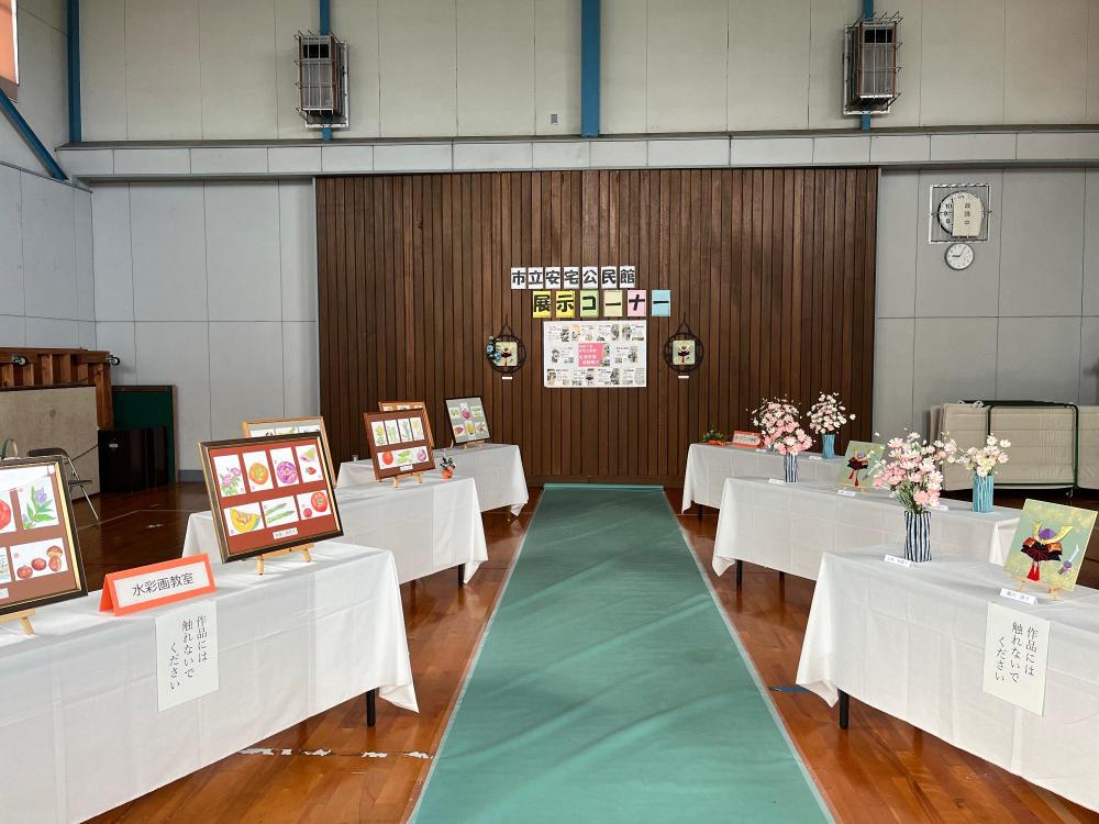 体育館に長い机が6本ありその上に公民館で作った作品が飾られている。花や額に入った絵葉書の作品など