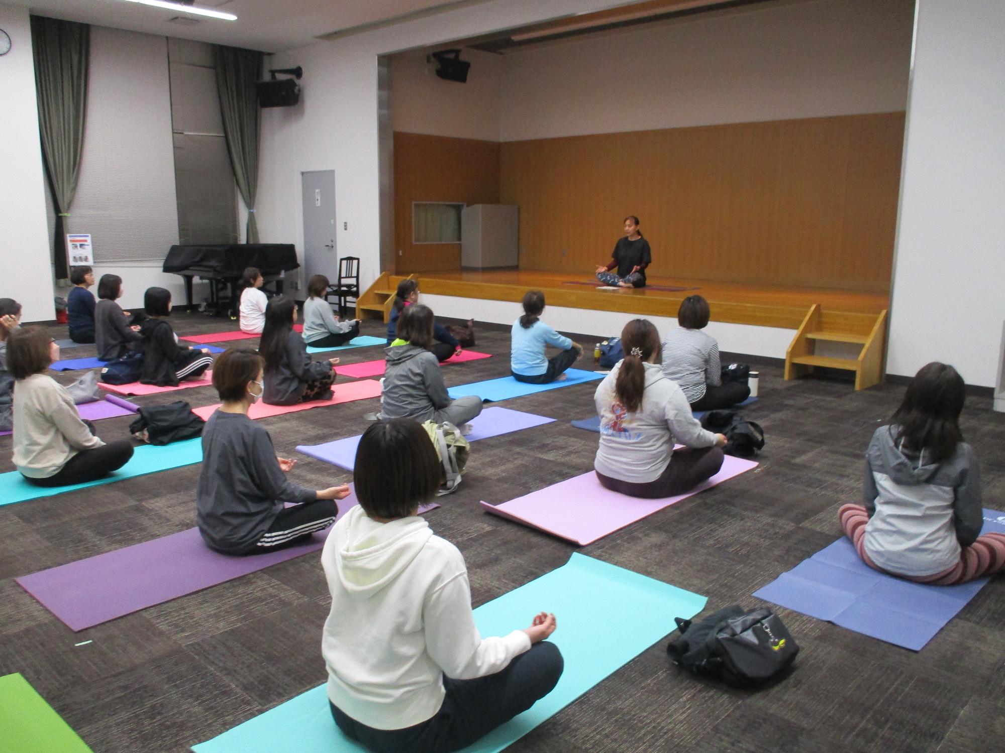 ヨガマットのうえにあぐらをかいて先生の説明を聞く様子
